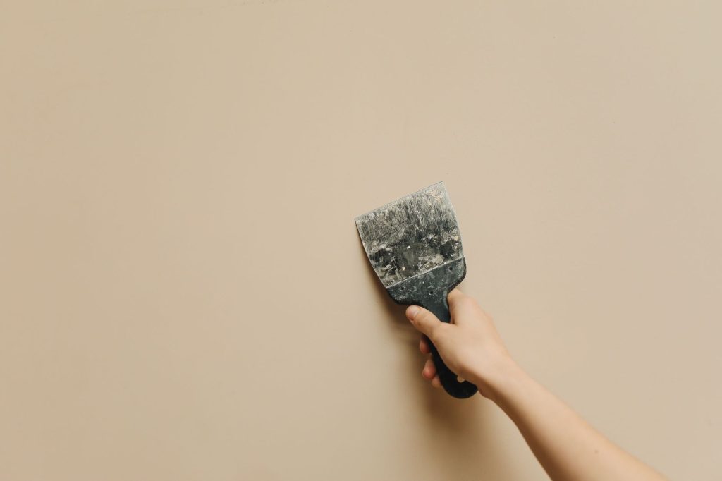 Hand hält einen gebrauchten Spachtel an eine glatte, beige Wand