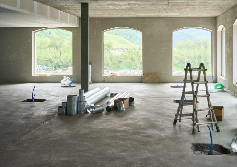 Wie lange trocknet Estrich? Richtwerte je Estrichart und Dicke Wie lange trocknet Estrich – Rohbau-Raum mit unbelegtem Boden und großen Fenstern